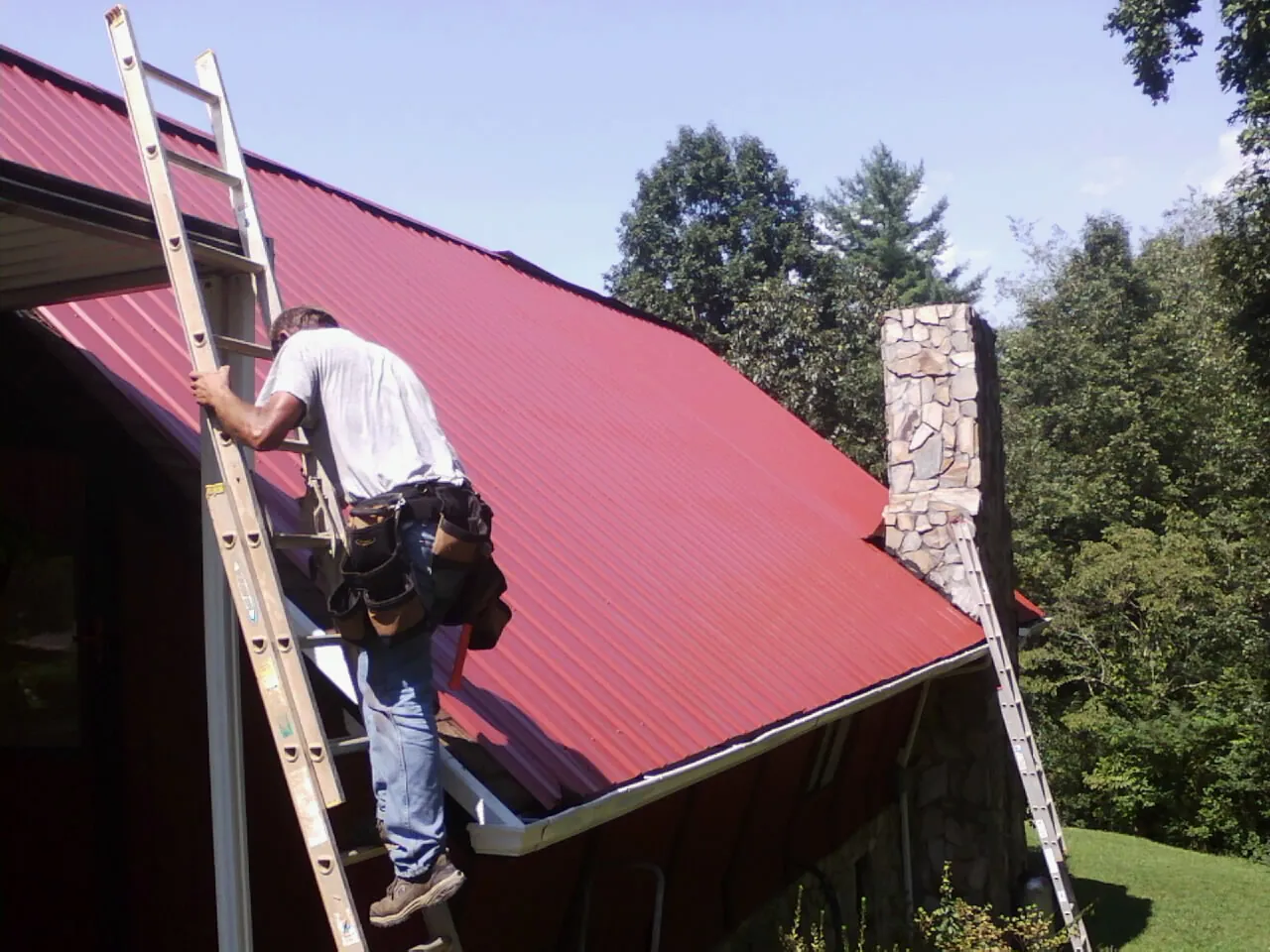 Roofer on a ladder beside a red metal roof and stone chimney — Soffit & Fascia Repair by Roofing Brothers in Glenville